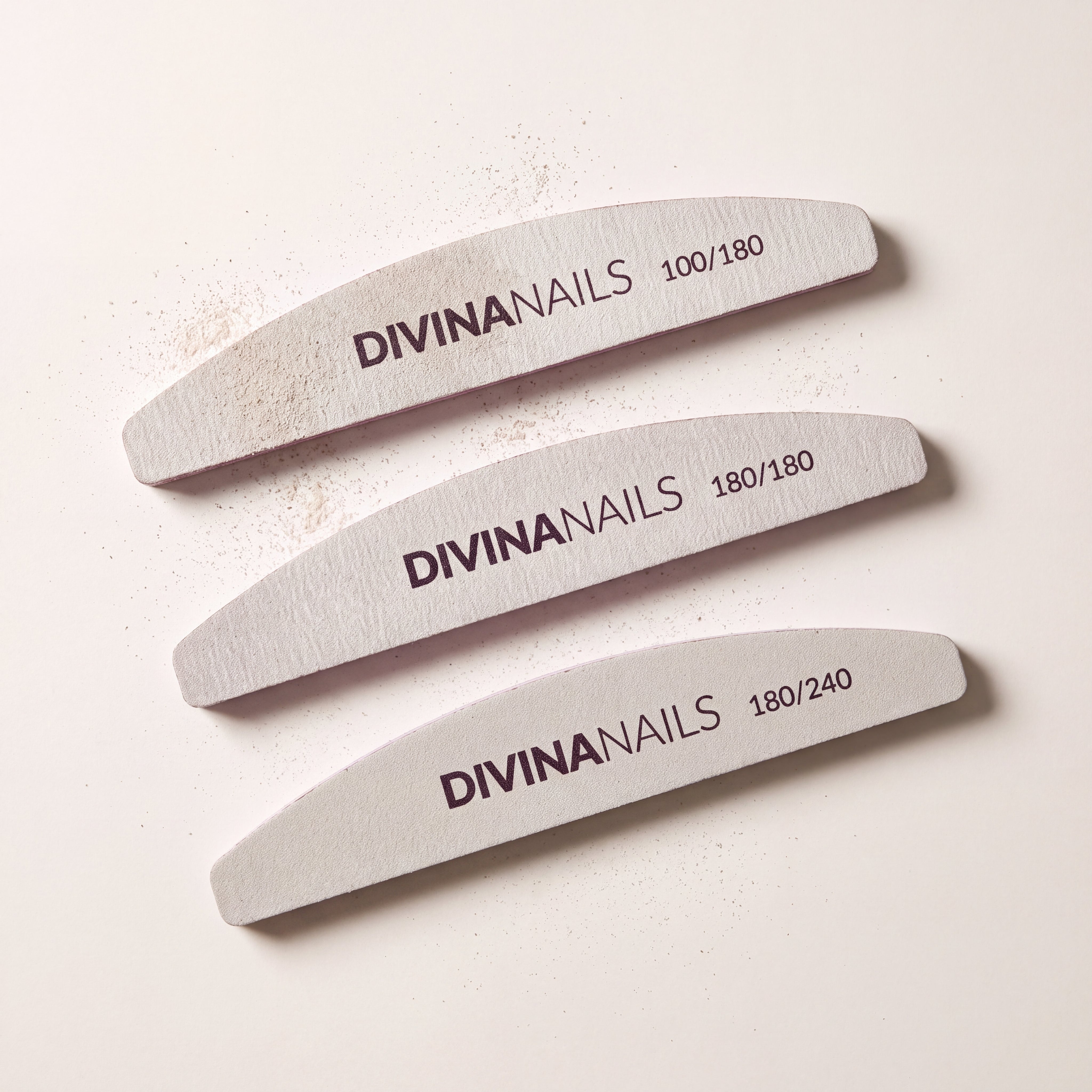 Tre lime mezzaluna Divina Nails di grana 100/180, 180/180 e 180/240 allineate dall'alto, dalla piu' grossa alla piu' fine, con la polvere di limatura